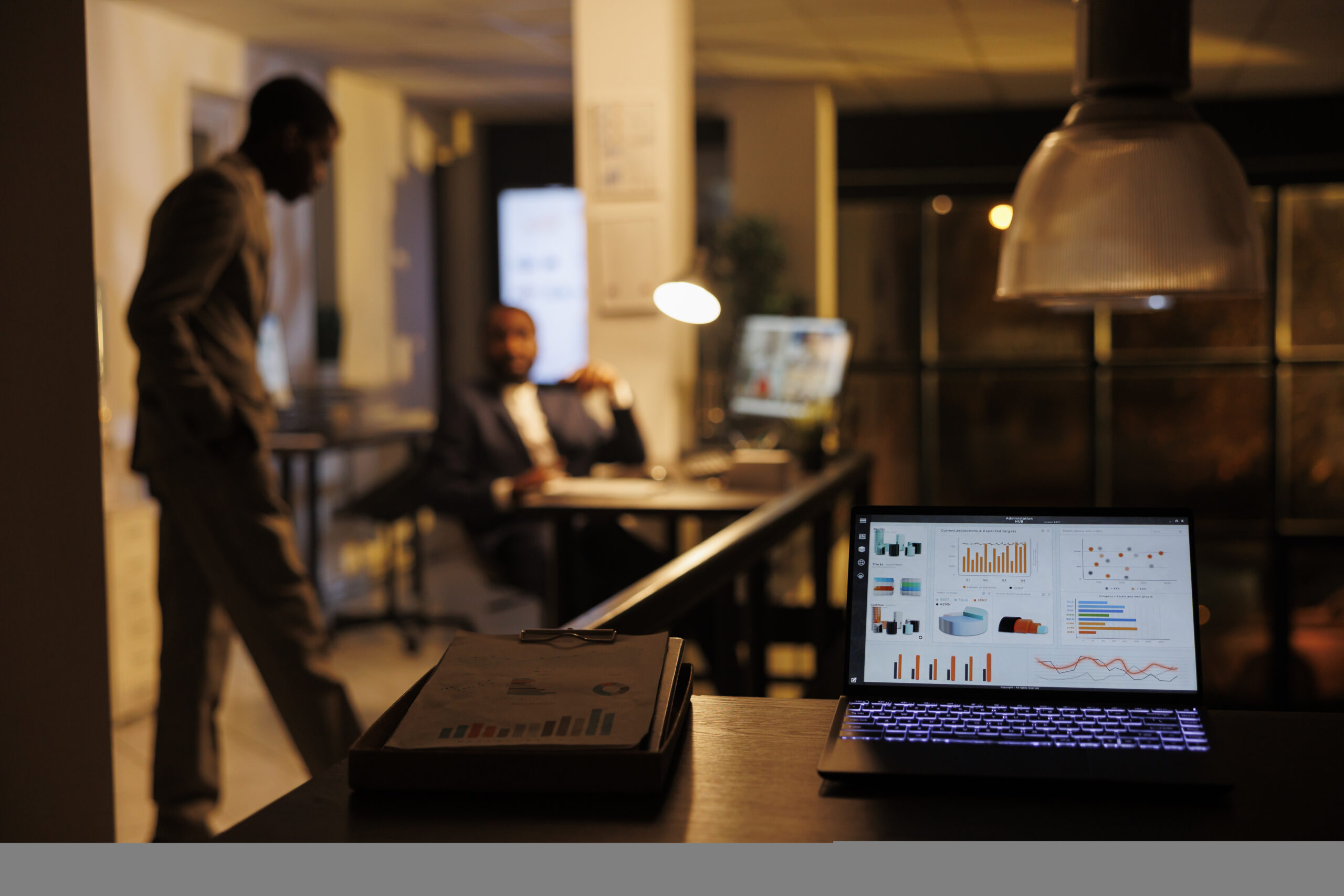 Laptop computer with company statistics standing on desk table in startup office, business team working overhours at marketing strategy in background. Coworkers discussing financial growth report